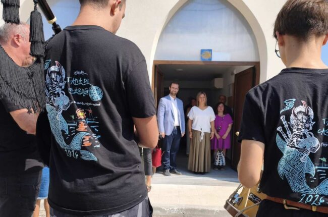 Two men in black T-shirts with blue graphic designs stand at the edge of a doorway, facing a group of guests gathered inside a light-colored building entrance.
